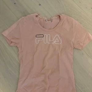 Ljusrosa FILA t-shirt i bomull - Snygg ljusrosa t-shirt från FILA med vit och svart logga framtill. Klassisk rund halsringning och korta ärmar. Tillverkad i mjuk bomull för en skön känsla. Perfekt för en avslappnad och sportig stil.