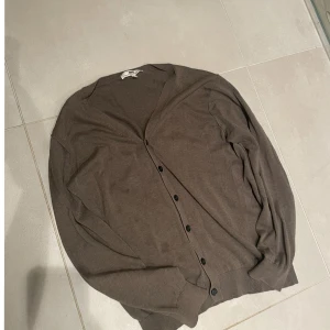 Brun kofta med knappar från H&M - Snygg brun kofta från hope med v-ringad hals och svarta knappar framtill. Koftan har långärmade ärmar och är gjord i mjuk bomull, perfekt för lager-på-lager. Enkel och stilren design som funkar till många olika looks.