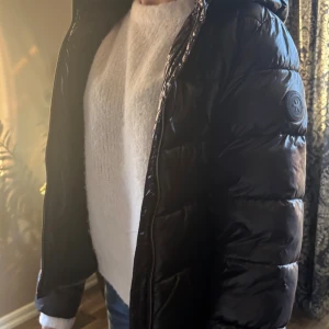 Svart pufferjacka från Everest - Svart pufferjacka från Everest med glansig yta och vadderade sektioner. Jackan har huva, lång ärm och dragkedja framtill. Märkets logga sitter på vänster ärm. Perfekt för kalla dagar och har en clean, modern look.