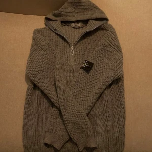 Stickad half zip från Stone Island - Säljer en grå stickad hoodie från Stone Island med klassisk patch på ärmen. Tröjan har huva, half zip och ribbade muddar. Perfekt för dig som gillar streetwear och vill ha något som sticker ut lite extra. Grym passform och riktigt skön att bära.