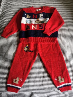 Julklappstips! Röd stickad jultröja och byxor med djurmotiv - Stl 86/92. Helt ny i sin förpackning. Supergullig röd stickad tröja med lång ärm och knappar vid axeln. Framsidan har broderade djurmotiv, text och färgglada detaljer i vitt, blått och grönt. Perfekt för julen med sina festliga inslag och mjuka material. Matchande byxor med söta djur och ränder vid benslut ingår. Kolla mina andra annonser för samfrakt. 