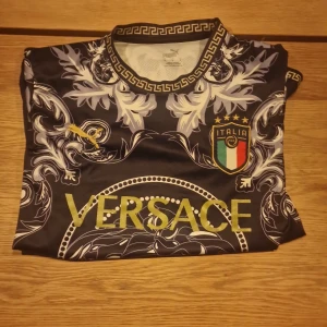 Italien Versace x Puma fotbollströja - Unik svart fotbollströja med Versace-tryck, guldiga detaljer och Italiens landslagsemblem. Tröjan har lång ärm, barockinspirerat mönster och rund hals med grekisk kant. Materialet är lätt och andas, perfekt för match eller streetwear.