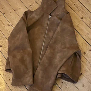 Läder jacka från H&M  - Nypris- 700kr Säljer en snygg brun bomberjacka i mocka-look med hög krage och silverfärgad dragkedja framtill. Jackan har en loose passform och är perfekt för dig som gillar stilrena och enkla plagg med lite edge. Passar till många olika outfits och har en modern vibe.