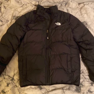 Svart pufferjacka från The North Face - Svart pufferjacka från The North Face med klassisk logga på bröstet och ryggen. Jackan har hög krage, två sidofickor och dragkedja framtill. Perfekt för kalla dagar med sin vadderade design och stilrena look. Jackan går att vända ut och in som ni ser på bilderna. L i junior storlek men passar XL i junior och kanske XXS vuxen. 