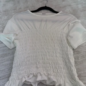 Vit smockad blus med volangkant - Supersöt vit blus med smockad midja och volangkant nertill. Blusen har korta ärmar och rund halsringning. Perfekt för dig som gillar en feminin och trendig stil.