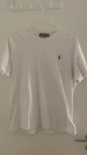 Vit t-shirt från Polo Ralph Lauren - Vit t-shirt från Polo Ralph Lauren med klassisk rund hals och korta ärmar. T-shirten är i mjuk bomull och har den ikoniska broderade loggan i marinblått på bröstet. Enkel och stilren design som funkar till allt.