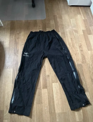 Svarta täckbyxor Arc'teryx - Svarta täckbyxor från Arc'teryx i storlek 3XL. Byxorna har elastisk midja med snörning, långa dragkedjor längs benen och logga på låret. Perfekta för utomhusaktiviteter och tillverkade i slitstarkt syntetmaterial.