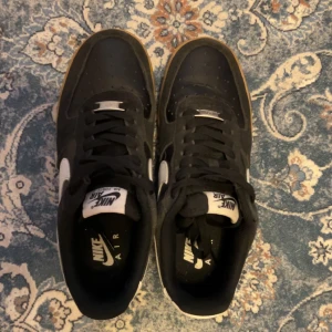 Nike Air Force 1 svart/gum sneakers - Svarta Nike Air Force 1 sneakers med vit swoosh och vit etikett på plösen. Skorna har en klassisk låg siluett och en snygg gummisula i brun/gul ton. Materialet är skinn och syntet, med snörning framtill. Perfekt för dig som gillar streetwear och tidlös stil.