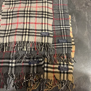 Burberrys rutig kashmirhalsduk - Klassisk halsduk från Burberrys i 100% kashmir med ikoniskt rutigt mönster. Finns i både grå och beige färg med svarta, vita och röda detaljer. Mjuk och lyxig känsla, med fransar i ändarna och Burberrys-märke. Perfekt accessoar för att lyfta din stil. 