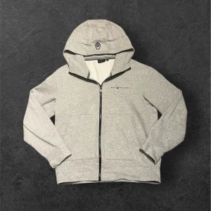 Grå hoodie från Sail Racing med dragkedja - Snygg grå hoodie från Sail Racing med svart dragkedja och huva. Tröjan har logga på bröstet och en mjuk insida. Perfekt för chill dagar och har två praktiska fickor framtill. Passformen är normal och materialet känns som bomull och polyester.