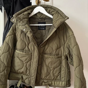 Olivgrön quiltad jacka från Zara - Quiltad jacka från Zara i olivgrön färg med coolt mönster och hög krage. Jackan har två stora fickor framtill, dragkedja och tryckknappar. Materialet är syntet och passformen är loose, perfekt för lager på lager under höst och vinter. Skriv vid frågor💗