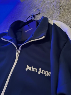 Blå track jacket från Palm Angels - Snygg marinblå track jacket från Palm Angels med vita ränder längs ärmarna och vit dragkedja, två fickor med dragkedja och Palm Angels-logga  på bröstet. Pris går att diskutera, är äkta. Tag medföljer