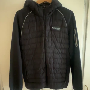 Svart pufferjacka från McKenzie - Svart pufferjacka från McKenzie med huva och dragkedja framtill. Jackan har quiltad framsida, släta ärmar och en diskret logga på bröstet. Snygga detaljer med reflexband längs axlarna och en ficka med dragkedja på ena ärmen. Perfekt för kyliga dagar.