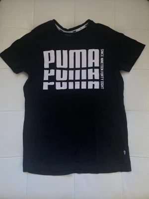 Svart Puma t-shirt med vit logga - Svart t-shirt från Puma med stor vit logga och texten 'Since Nineteen Forty Eight' på framsidan. Klassisk rund halsringning och korta ärmar. Tillverkad i mjuk bomull, perfekt för en sportig och avslappnad stil.