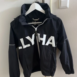 Svart vindjacka Helly Hansen 164/14 - Svart vindjacka från Helly Hansen med stor vit logga över bröst och rygg. Jackan har huva, hel dragkedja framtill och elastiska muddar vid ärmslut. Snygg och sportig look, perfekt för blåsiga dagar. Tillverkad i lätt polyester.