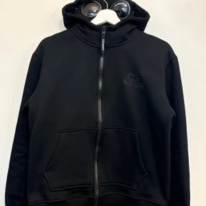  CP Company Hoodie - Svart hoodie från CP Company med ikonisk huva som har inbyggda goggles. Tröjan har dragkedja framtill, ribbade muddar och broderad logga på bröstet. Perfekt för dig som gillar streetwear och vill sticka ut med en unik detalj på huvan.