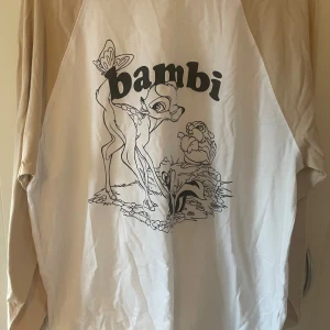 Beige och vit Bambi-tröja från H&M - Säljer en beige och vit långärmad tröja från H&M Divided med Bambi-tryck framtill. Tröjan har raglanärmar i beige och kroppen i vitt, med svart Bambi-motiv och text. Mjuk bomullskvalitet och normal passform. Perfekt för dig som gillar Disney och chill stil.