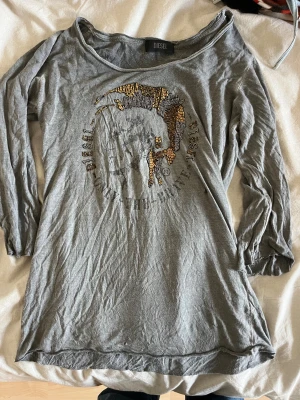 Grå långärmad tröja från Diesel - Snygg grå långärmad tröja från Diesel med bred halsringning och coolt paljettbroderat motiv framtill i guld och silver. Tröjan är i mjuk bomull och har ett avslappnat, lite loose fit. Perfekt för dig som gillar statement-plagg och streetstyle.