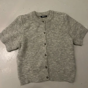 Grå stickad kofta från Gina Tricot - Kortärmad grå stickad kofta från Gina Tricot med rund hals och ribbade muddar. Koftan har knappar framtill i metall och är gjord i mjukt material som känns skönt mot huden. Perfekt för lager-på-lager och chill stil. Knappt använd