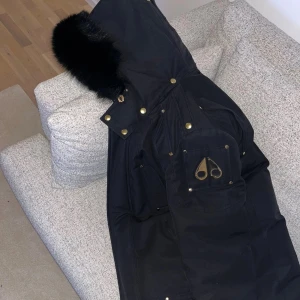 Svart Moose Knuckles med äkta päls - Säljer en svart dunjacka från Moose Knuckles med lyxig svart äkta päls på huvan och guldfärgade detaljer. Helt äkta ny pris går för 17000kr. Insidan är quiltad i guld med logomönster och broderad rygg. Jackan har dragkedja, knappar och flera fickor med ikonisk logga.