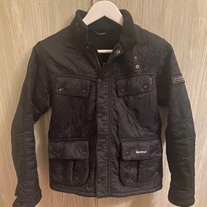 Svart quiltad jacka från Barbour stl 164 - Quiltad svart jacka från Barbour International med fyra fickor framtill, tryckknappar och dragkedja. Jackan har hög krage med mjukt foder och Barbour-logga på ärmen. Snygg, klassisk look med rutmönster och detaljer i metall. Storlek 164
