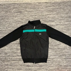 Svart kofta från Lacoste M - Svart kofta från Lacoste i storlek M med grönt rand över bröstet och klassisk krokodillogga. Koftan har hel dragkedja, ståkrage och broderad text 'Lacoste 1927' på ryggen. Tillverkad i lätt fleece, perfekt för en sportig och clean look.