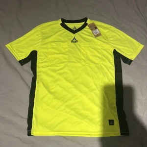 Gul domartröja från Select, S - Ordinarie pris 449. Säljer en gul domartröja från Select i storlek small. T-shirten har svarta detaljer längs sidorna och vid kragen, två bröstfickor och en sportig v-ringad design. Materialet är lätt polyester som andas, perfekt för fotbollsmatcher. 