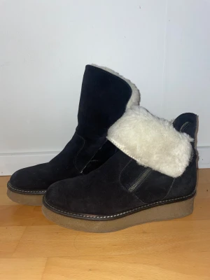 Svarta Shepherd boots med ullfoder - Mysiga svarta boots från Shepherd i mocka med fluffigt vitt ullfoder. Snyggt uppvik vid skaftet och grov platt sula i brunt. Dragkedja på sidan. Fodret har lossnat något vid hälen (se bild).