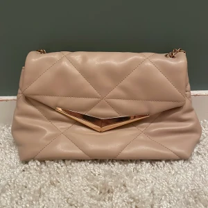 Beige quiltad axelväska med guldkedja - Snygg beige axelväska i quiltad design med guldfärgad kedja och coolt V-format spänne framtill. Väskan har ett stort fack och innerficka med dragkedja. Perfekt för dig som gillar stilrena detaljer och mjukt material.