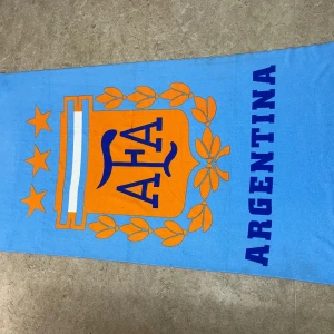 Argentina halsduk med AFA-logga - Snygg halsduk i ljusblått med stor orange AFA-logga och texten ARGENTINA i mörkblått. Dekorerad med stjärnor och bladmotiv, perfekt för dig som älskar fotboll och vill visa ditt stöd för Argentina. Rektangulär form och mjukt material.