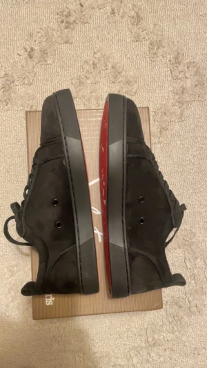 Svarta sneakers från Christian Louboutin - Säljer ett par svarta sneakers från Christian Louboutin med klassisk röd sula och snygg snörning. Skorna är tillverkade i mocka med rund tå och platt sula. Insidan är beige och har guldfärgad logga. Perfekt för dig som gillar exklusiv stil och detaljer.