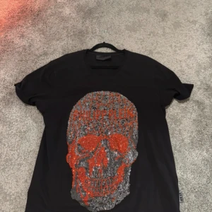 Svart Philipp Plein t-shirt med döskalle - Svart t-shirt från Philipp Plein i 100% bomull med stor glittrig döskalle framtill i orange och silver. Klassisk passform med korta ärmar och rund hals. Klassisk Philipp Plein logga i nacken. Pris går att diskutera.