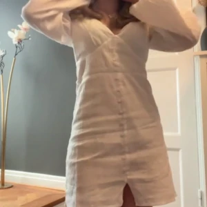 Vit kortklänning med v-ringning - Säljer en vit kortklänning med v-ringad hals och långa ärmar. Klänningen har knappar framtill och en liten slits nedtill. Materialet känns lätt och fräscht, perfekt för varmare dagar. Enkel och stilren design som är lätt att matcha.