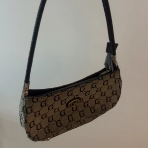 Beige axelväska från Guess - Snygg beige axelväska från Guess med klassiskt G-mönster över hela väskan och svarta detaljer. Väskan har guldfärgad logga framtill och svart axelrem. Perfekt storlek för det viktigaste och en stilren look.
