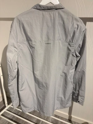 Ljusgrå overshirt från H&M - Snygg ljusgrå overshirt från H&M med två stora bröstfickor och svart dragkedja framtill. Skjortan har klassisk krage och långa ärmar med knapp vid manschetten. Perfekt att slänga på sig över en t-shirt för en chill look.