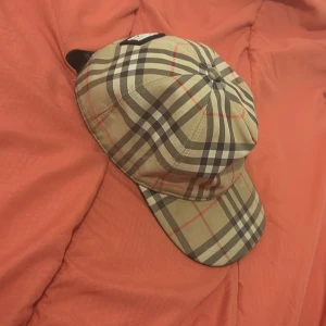 Burberry rutig keps i beige och svart - Säljer en klassisk Burberry keps i den ikoniska rutiga designen med beige, svart och röda detaljer. Kepsen har justerbar rem bak och en patch med texten 'Burberry London England'. Tillverkad i bomull för en skön och stilren look.