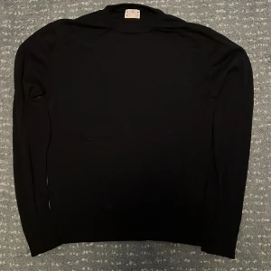 Mörkblå stickad tröja från ASKET - Mörkblå stickad tröja från ASKET med stilren design och långärmade smala ärmar. Tröjan har en hög krage och är tillverkad i mjukt material som känns skönt mot huden. Perfekt för dig som gillar minimalistisk och clean stil.