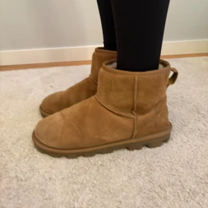Bruna UGG boots i mocka - Säljer ett par klassiska bruna UGG boots med rund tå och platt sula. Bootsens utsida är i mjuk mocka och insidan är fodrad med fluffigt material för extra värme. De har en låg siluett och praktisk hälflik baktill. Perfekta för kyliga dagar och jättesnygga vinterskor!!