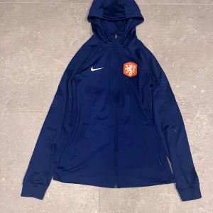 Blå Nike fotbollsjacka - Blå sportig jacka från Nike med huva och dragkedja. Jackan har KNVB-logga på bröstet och är tillverkad i ett lätt, syntetiskt Dri-Fit-material. Slim fit-modell med långa ärmar och två fickor framtill.
