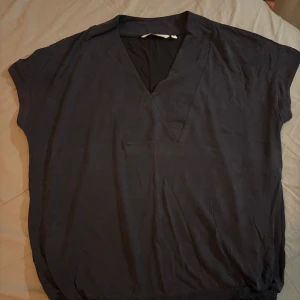 Svart blus från House of Lola - Svart blus från House of Lola i storlek L. Modellen har v-ringning, korta ärmar och en avslappnad passform med resår nertill. Perfekt för dig som gillar en enkel och stilren look. Tillverkad i mjuk modal/polyester som känns skön mot huden.