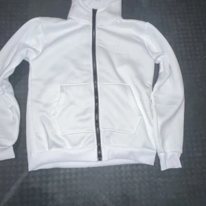 Vit hoodie från C.P. Company - Snygg vit hoodie från C.P. Company med broderad logga på bröstet. Tröjan har huva, hel dragkedja framtill och en stor magficka. Materialet känns mjukt och har en clean look som passar till streetwear. Perfekt för dig som gillar stilrena och bekväma plagg.