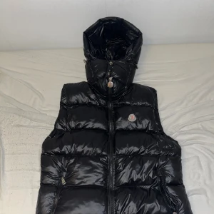 Svart dunväst från Moncler med huva - Svart, glansig dunväst från Moncler med avtagbar huva och ikonisk logga på bröstet. Västen har quiltad design, två sidofickor med dragkedja och en röd innerficka. Perfekt för lager-på-lager och streetstyle.