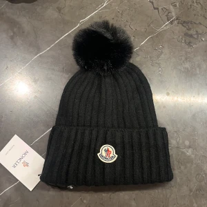 Svart Moncler mössa med pälsboll - Ribbstickad svart mössa från Moncler med stor fluffig pälsboll på toppen och klassisk Moncler-logga framtill. Perfekt för kalla dagar och ger en lyxig vibe till din vinterstil.