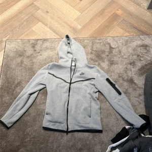 Grå hoodie från Nike med dragkedja - Snygg grå hoodie från Nike med hel dragkedja och huva. Tröjan har lång ärm, fickor fram och en extra ficka med dragkedja på ena ärmen. Perfekt för chill eller träning, stilren logga på bröstet.