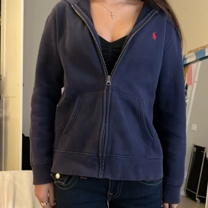 Marinblå hoodie från Ralph Lauren - Säljer en marinblå hoodie från Ralph Lauren med dragkedja och huva. Tröjan har två stora fickor framtill och den klassiska röda Polo-loggan broderad på bröstet. Perfekt för chill dagar och enkel att matcha med jeans.