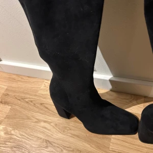 Svarta mockastövlar med klack - Snygga svarta stövlar i mocka med bred klack och rund tå. Stövlarna går upp över knäet och har en diskret dragkedja på insidan för enkel påtagning. Perfekta för att lyfta din outfit med en stilren och modern look.