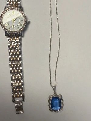 Silverfärgad Geneva klocka med strass - Stilren klocka från Geneva med rund urtavla i silver och guldton, romerska siffror och gnistrande strass runt boetten. Länken är tvåfärgad i silver och guld, vilket ger en lyxig känsla. Perfekt accessoar för dig som gillar bling och klassisk design.Ny Geneva klocka och en silver halsband stämplat 