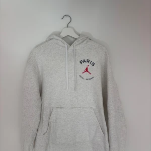 Grå Jordan Paris Saint-Germain hoodie - Snygg ljusgrå hoodie från Jordan x Paris Saint-Germain med röd Jumpman-logga och text på bröstet. Stor Paris-tryck och siffran 23 i rött på ryggen. Klassisk känguruficka, huva med snörning och mjukt material. 