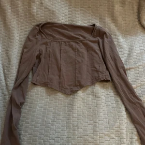 Brun croppad långärmad korsett-topp - Säljer en brun croppad korsett-topp med långa ärmar och markerade sömmar framtill. Toppen har en snygg fyrkantig halsringning och är tillverkad i mjukt bomullsmaterial. Perfekt för dig som gillar trendiga och stilrena plagg med lite edge.