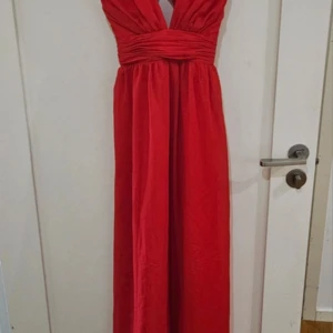 Röd långklänning med djup v-ringning - Snygg röd långklänning från Nelly med djup v-ringning och korsade axelband i ryggen. Klänningen har markerad midja med rynkade detaljer och är ärmlös. Perfekt till balen!